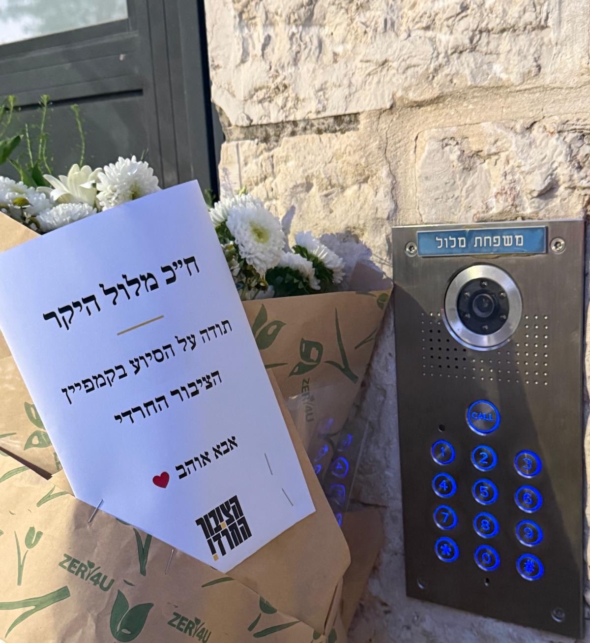 זר פרחים לבן וכרטיס ברכה שנשלחו לביתו של חבר הכנסת ארז מלול, לצד אינטרקום שעליו שלט «משפחת מלול»